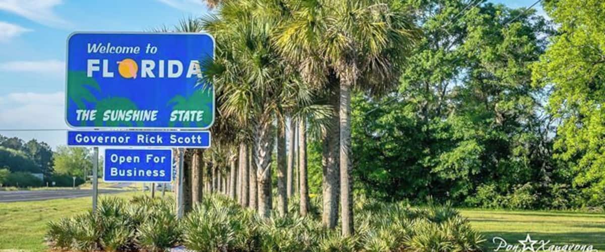 A welcome sign to Florida with palm trees and a clear sky.