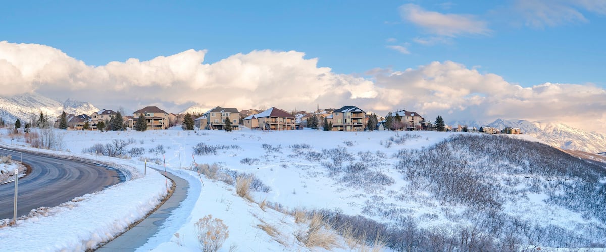 A snow-covered residential area with houses on a hillside, a winding road, and snow-covered mountains in the background.