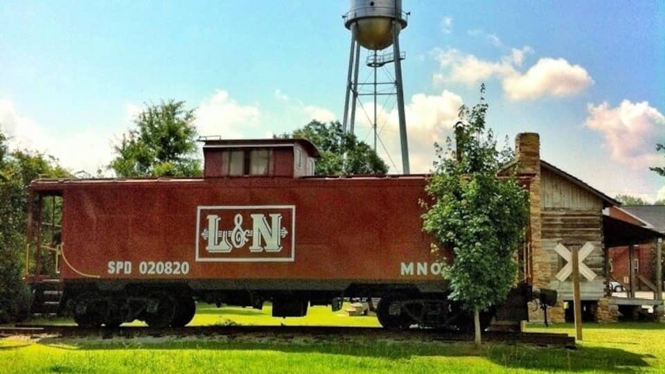 A red caboose with 'L&N' and 'SPD 020820' on it, a water tower, and a wooden building with a crossbuck sign.