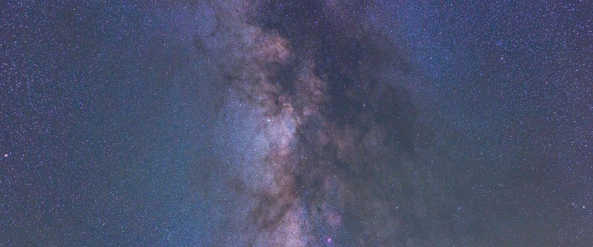 A night sky with the Milky Way over a calm body of water and a rocky shoreline with wooden posts.