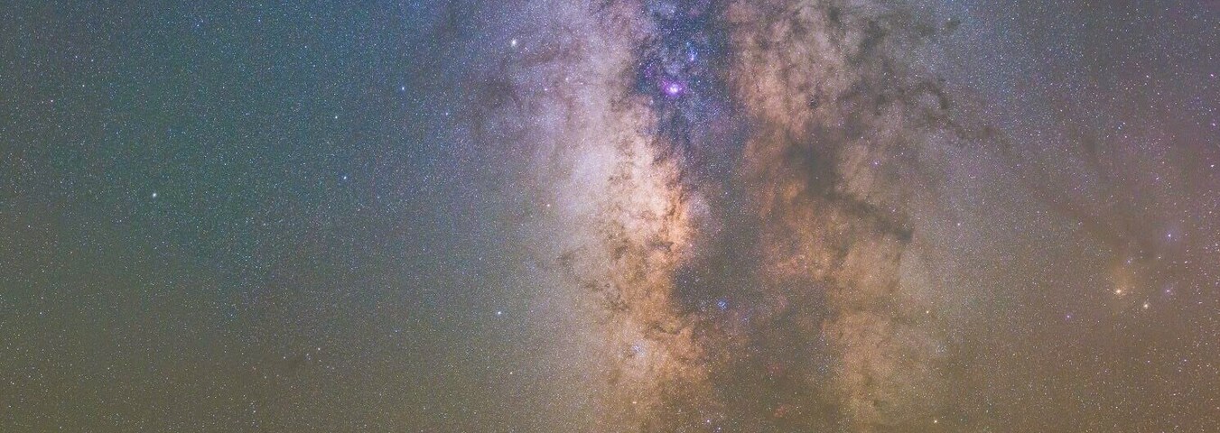 A night sky with the Milky Way over a calm body of water and a rocky shoreline with wooden posts.