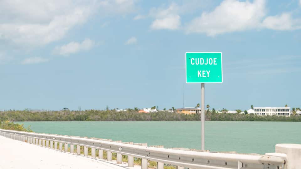 A sign reading "CUDJOE KEY" by a coastal road with a clear sky and calm sea.