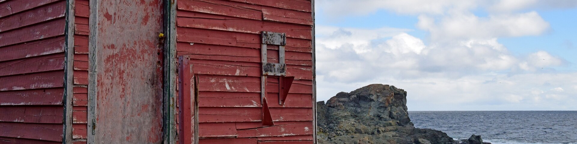 Una cabaña de madera roja junto al mar, con un promontorio rocoso al fondo.