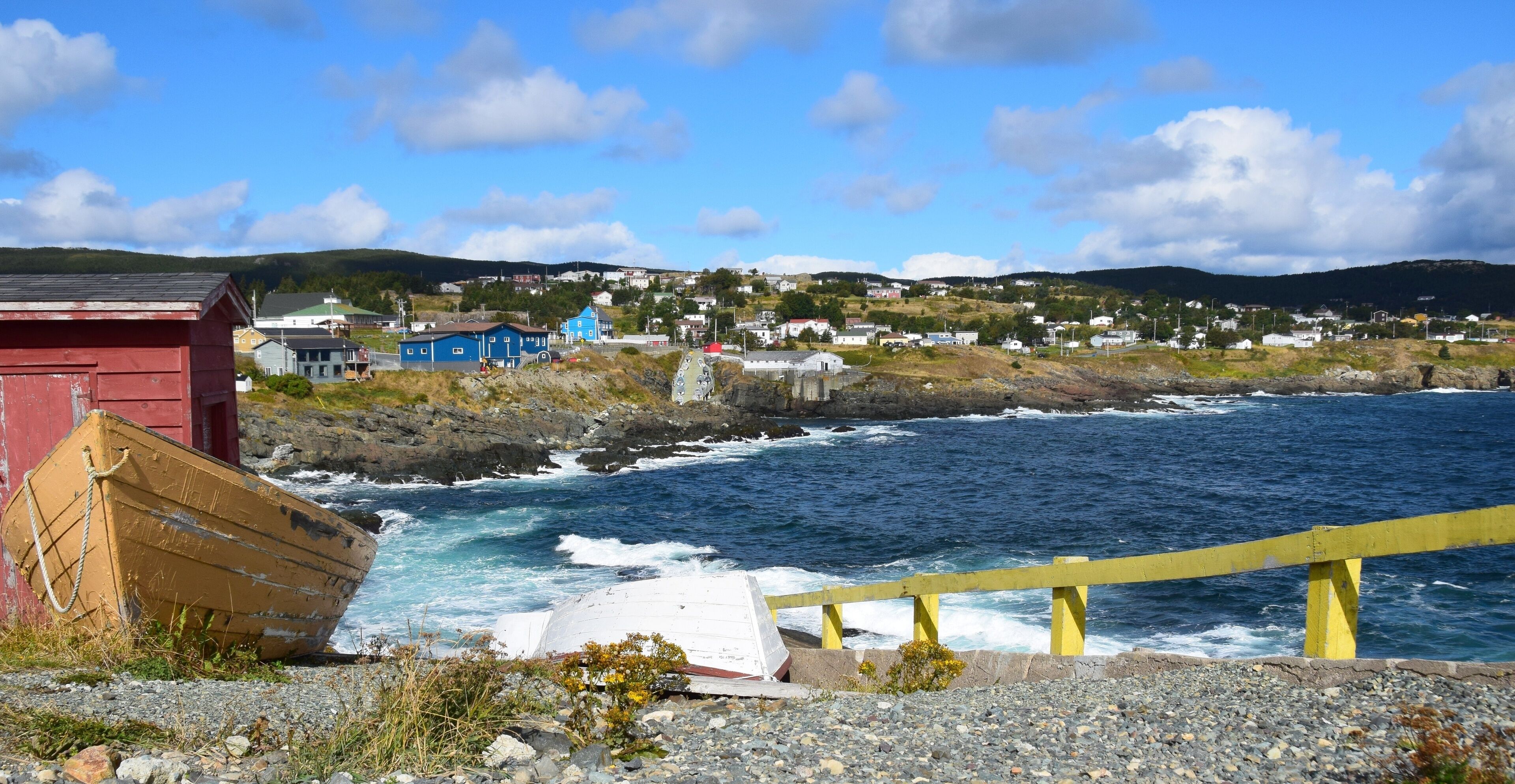 A coastal scene with a rocky shoreline, a wooden boat, a yellow railing, and a village in the background.