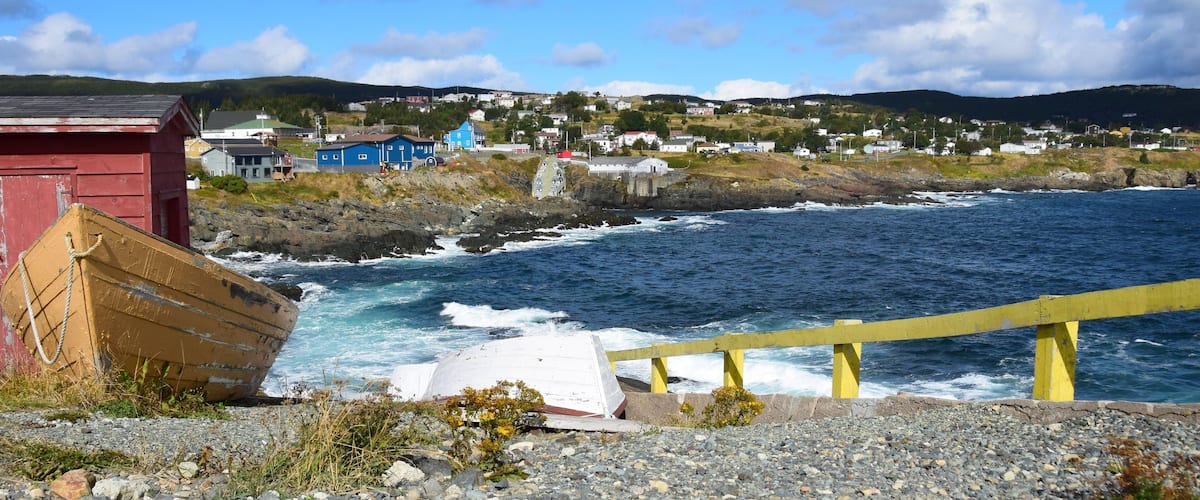 A coastal scene with a rocky shoreline, a wooden boat, a yellow railing, and a village in the background.