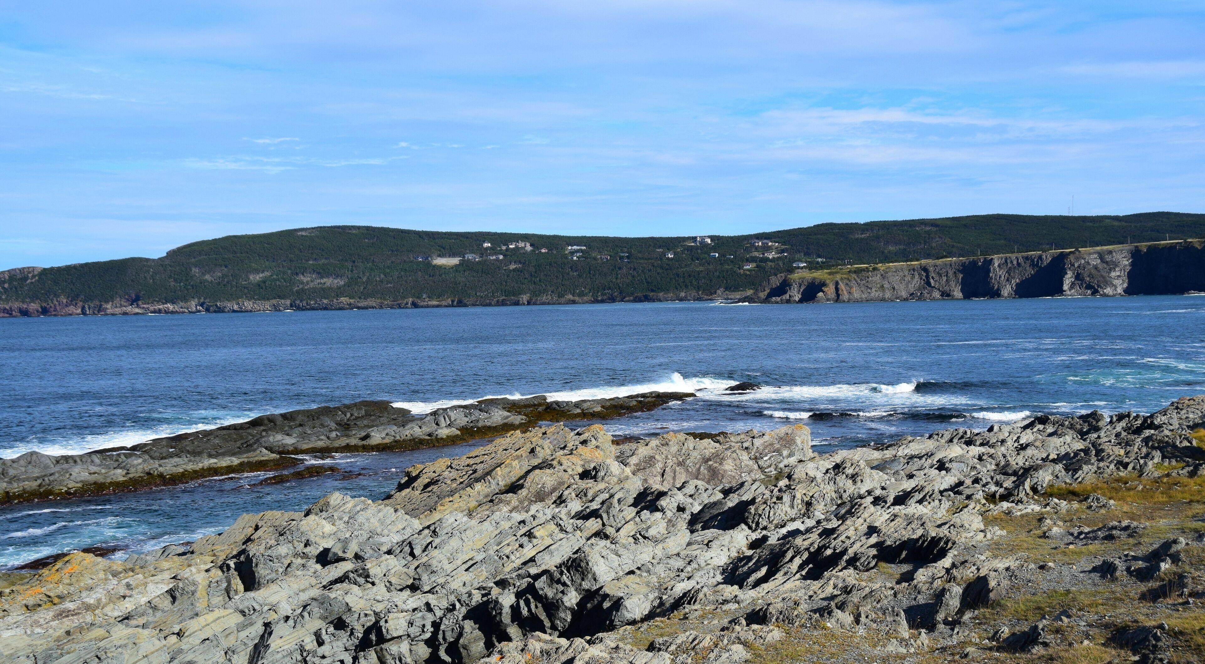 A rocky coastline with waves crashing against the shore, a clear blue sky, and a distant view of a forested hill with buildings.