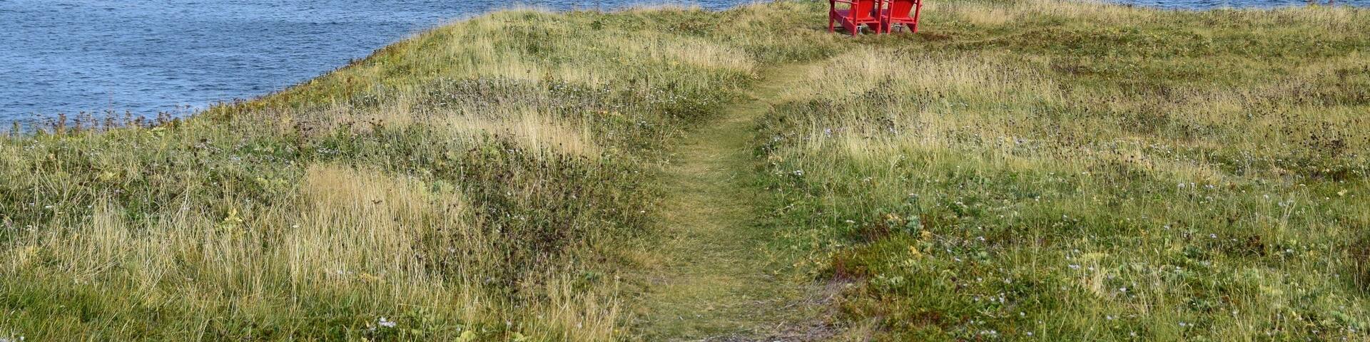 two empty red Adirondack chairs on the edge of a cliff overlooking the ocean and coastline, Silver Mine Head Path Torbay Newfoundland Canada