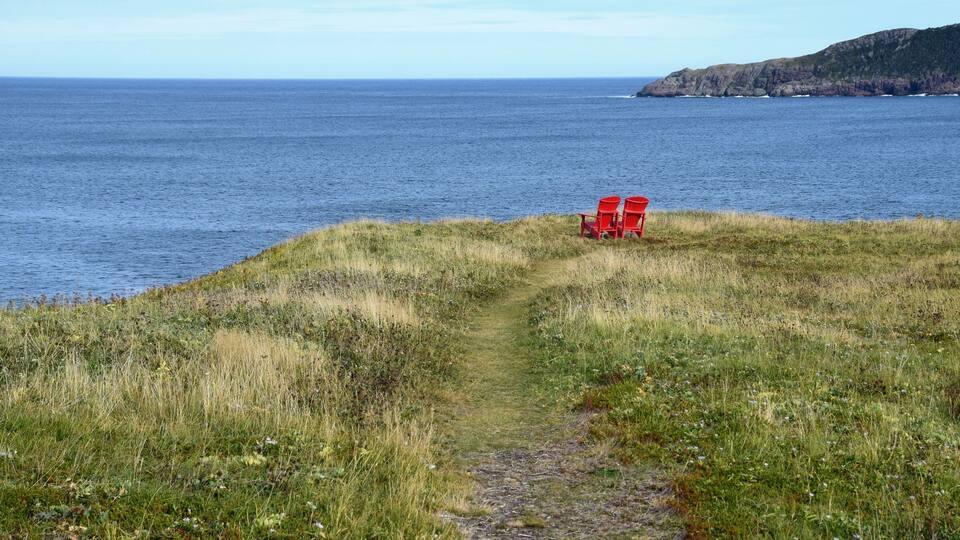 two empty red Adirondack chairs on the edge of a cliff overlooking the ocean and coastline, Silver Mine Head Path Torbay Newfoundland Canada