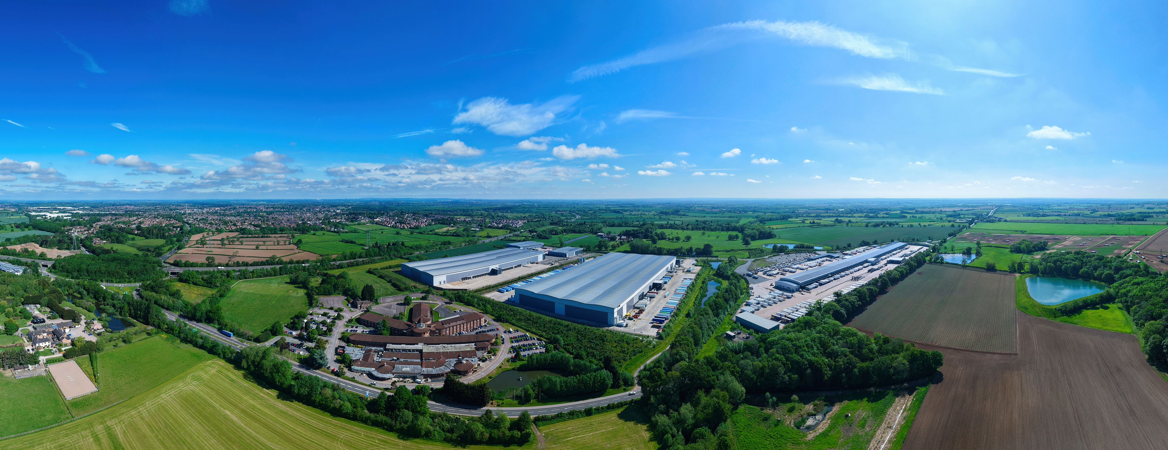 An aerial view of a large industrial area with multiple warehouses, surrounded by green fields and a small lake.