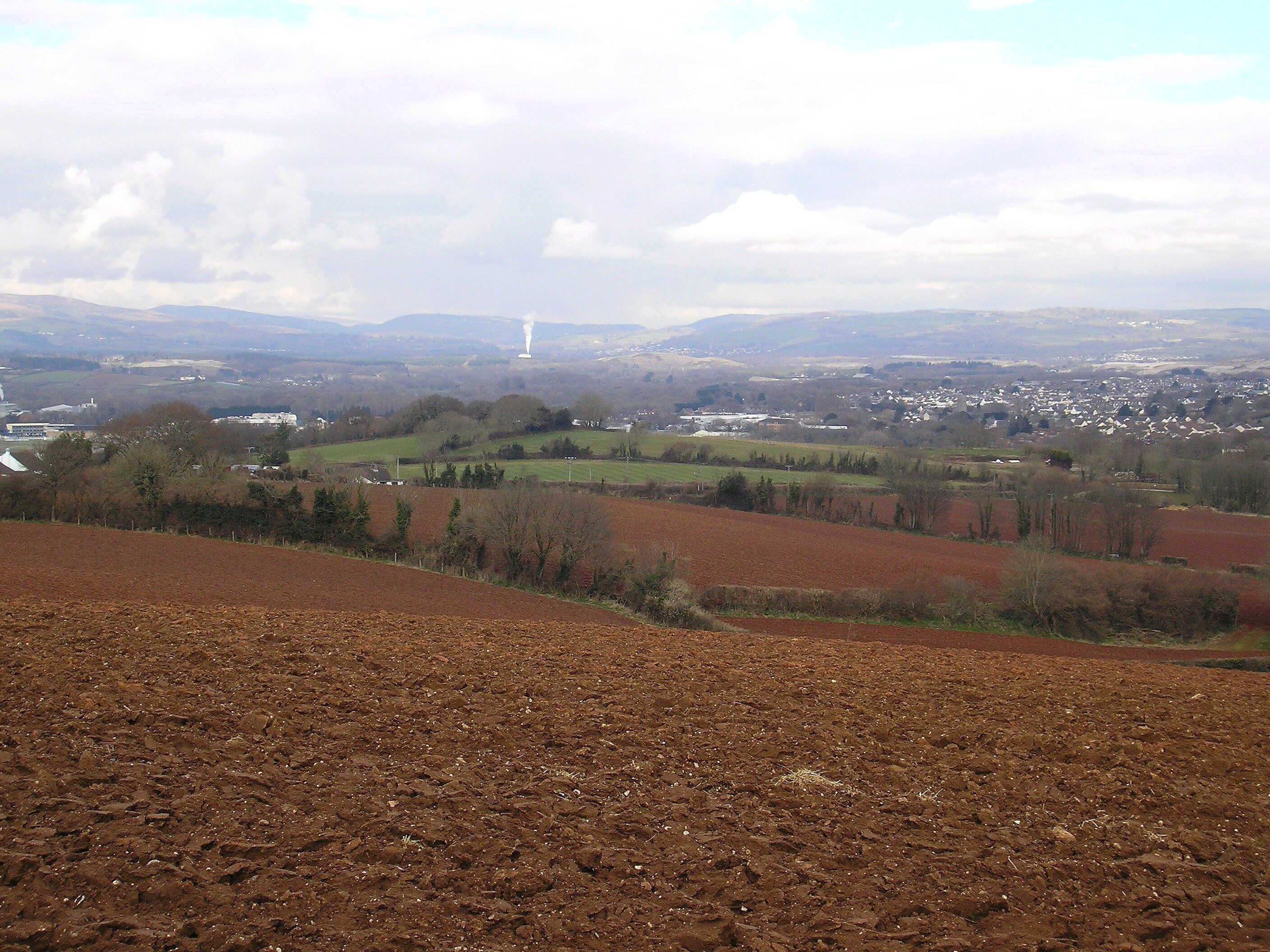 A rural landscape with plowed fields, scattered trees, and distant buildings.