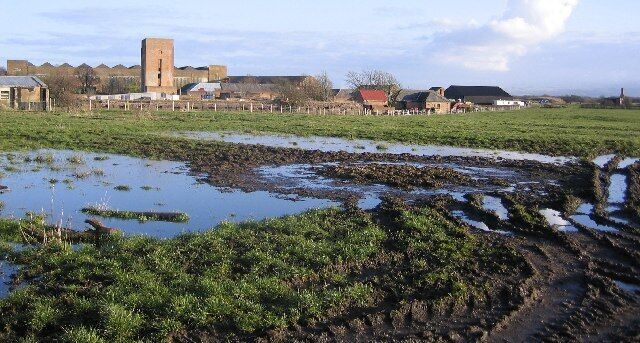A rural landscape with a large open field, patches of water, and a distant building with a tower.