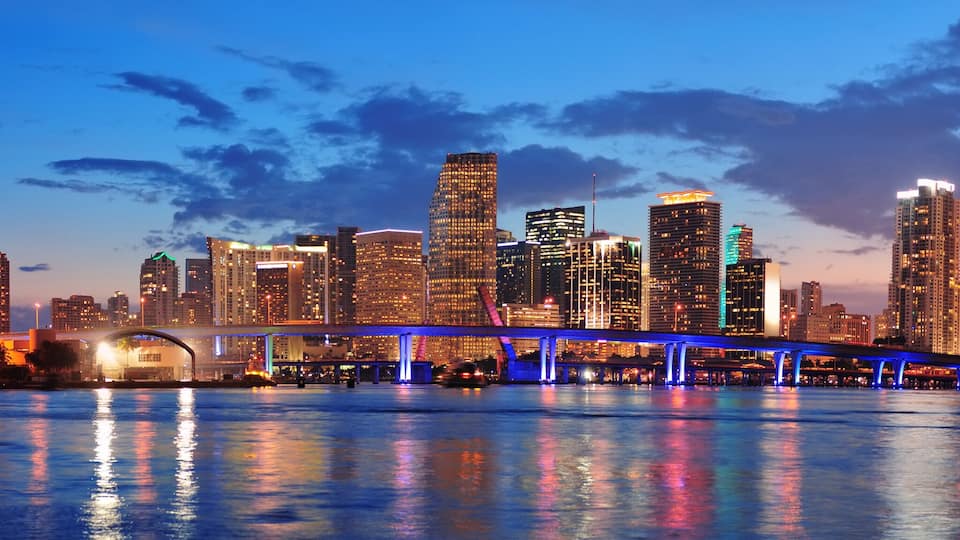 Linha do horizonte urbana com edifícios iluminados e uma ponte com iluminação azul sobre a água.