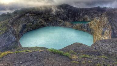 A volcanic crater with a turquoise lake surrounded by rugged terrain and a cloudy sky.