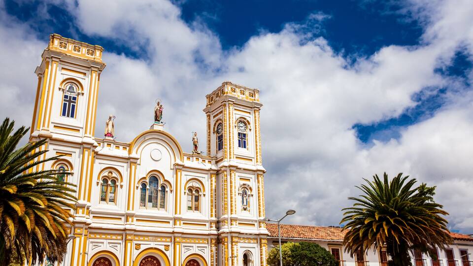 A historic church with two towers and statues, surrounded by palm trees and under a partly cloudy sky.