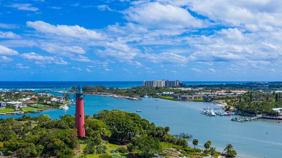 A coastal town with a prominent red lighthouse, a marina with boats, and a backdrop of buildings and greenery.