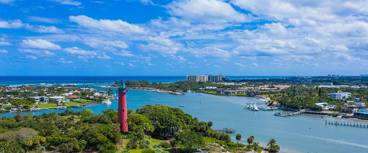 A coastal town with a prominent red lighthouse, a marina with boats, and a backdrop of buildings and greenery.