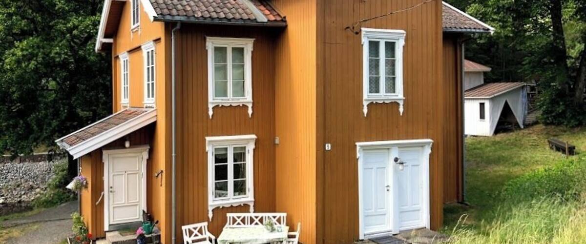 A two-story wooden house with a brown exterior and white trim, a front porch with a bench, and a small garden.