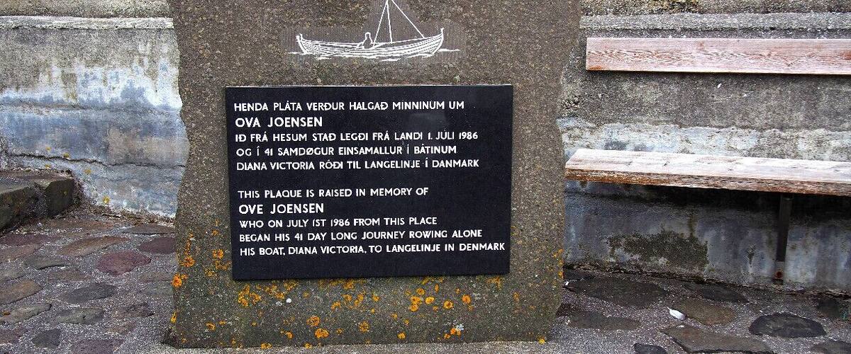 A memorial plaque with a boat illustration and text commemorating Ove Jensen's 1986 rowing journey from this location to Denmark.