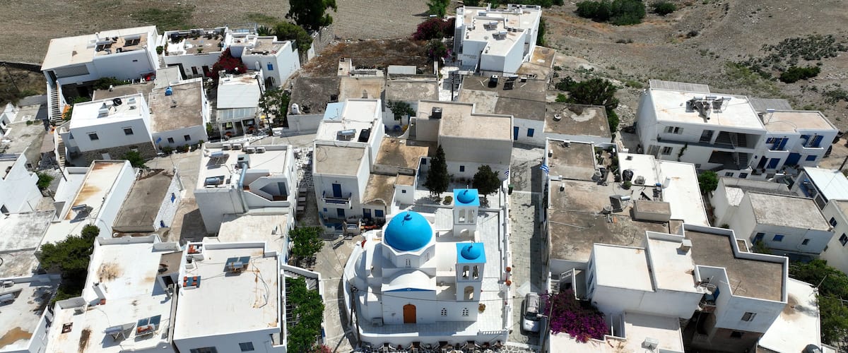 An aerial view of a white-washed village with blue domes, narrow streets, and rocky terrain.