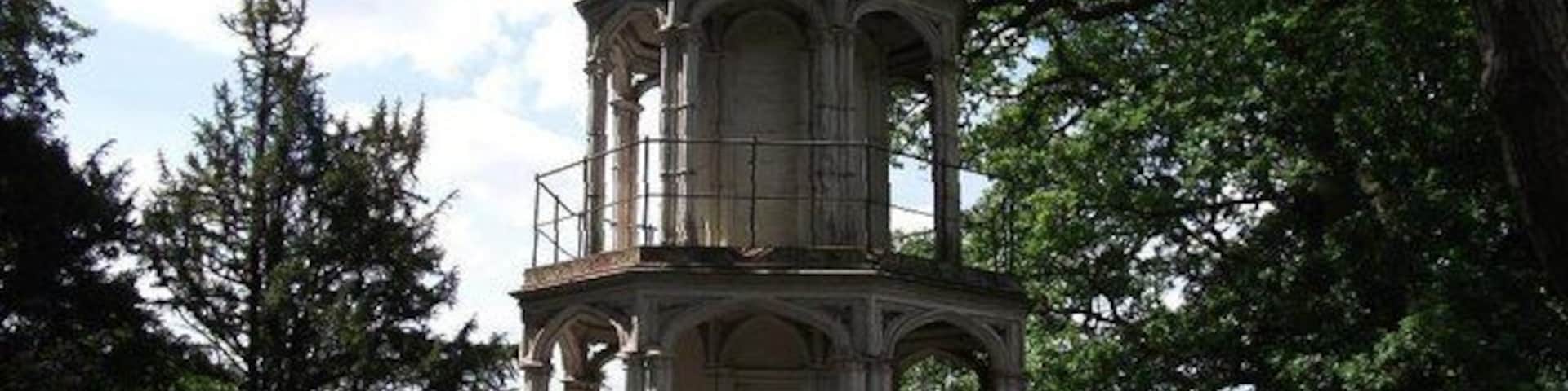 Uma torre histórica com relógio, cercada por árvores e um caminho.