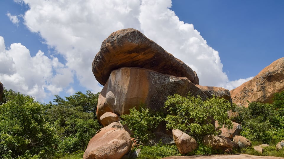 A large rock formation with a balanced boulder atop, surrounded by greenery and under a clear blue sky.