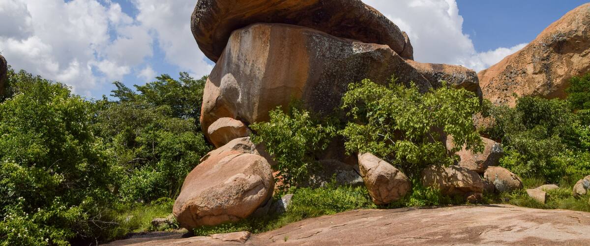 A large rock formation with a balanced boulder atop, surrounded by greenery and under a clear blue sky.