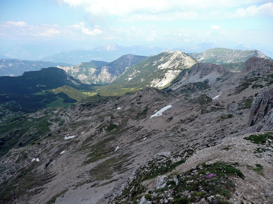 A rugged mountain landscape with rocky terrain, patches of snow, and distant mountain ranges under a clear sky.