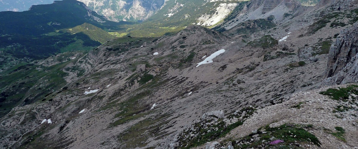A rugged mountain landscape with rocky terrain, patches of snow, and distant mountain ranges under a clear sky.