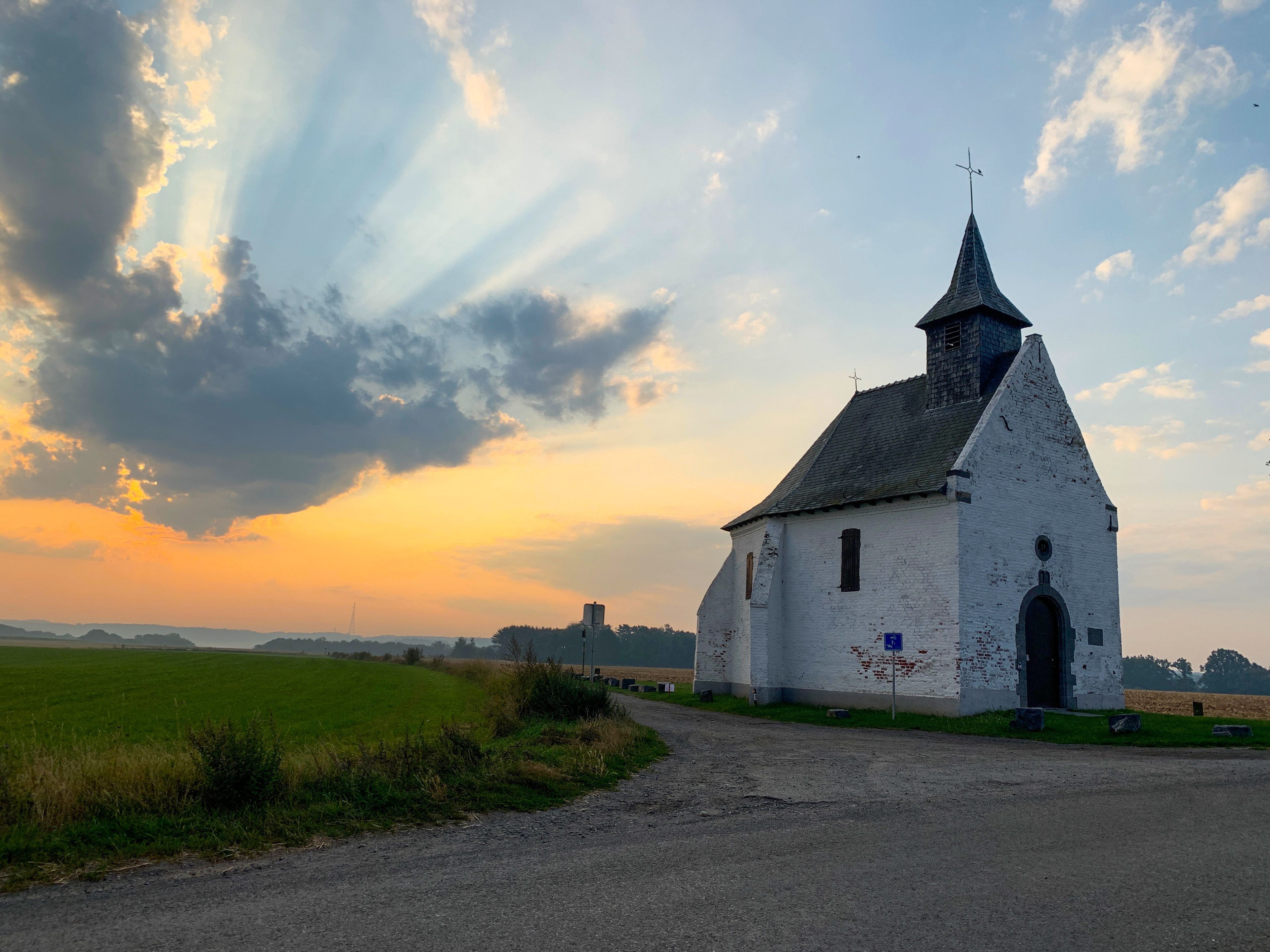 Een witte kerk met een puntige klokkentoren en een kruis bovenop, omgeven door een landelijk landschap met velden en een onverharde weg.