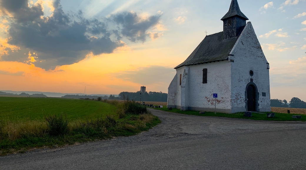 Een witte kerk met een puntige klokkentoren en een kruis bovenop, omgeven door een landelijk landschap met velden en een onverharde weg.