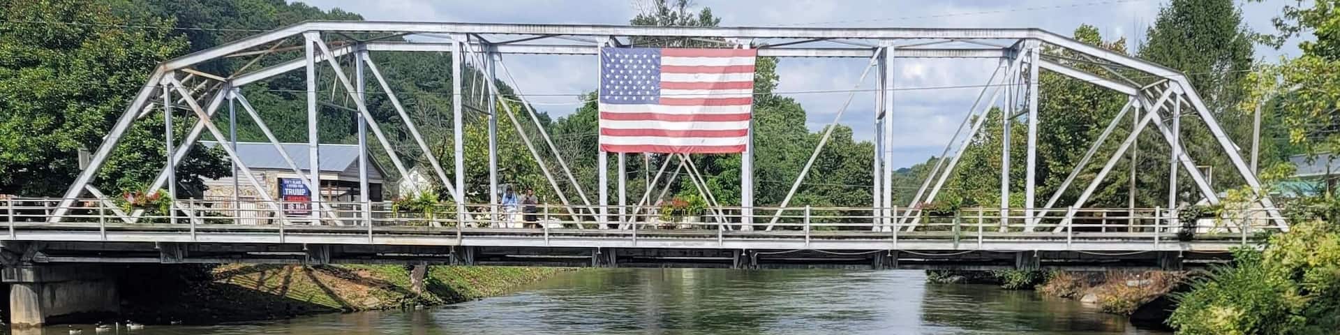 A bridge with an American flag, a river, and greenery in the background.