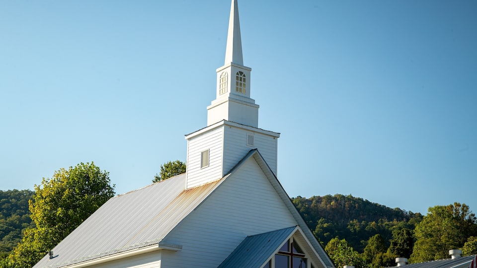 A white church with a steeple and a blue roof, surrounded by trees and hills.