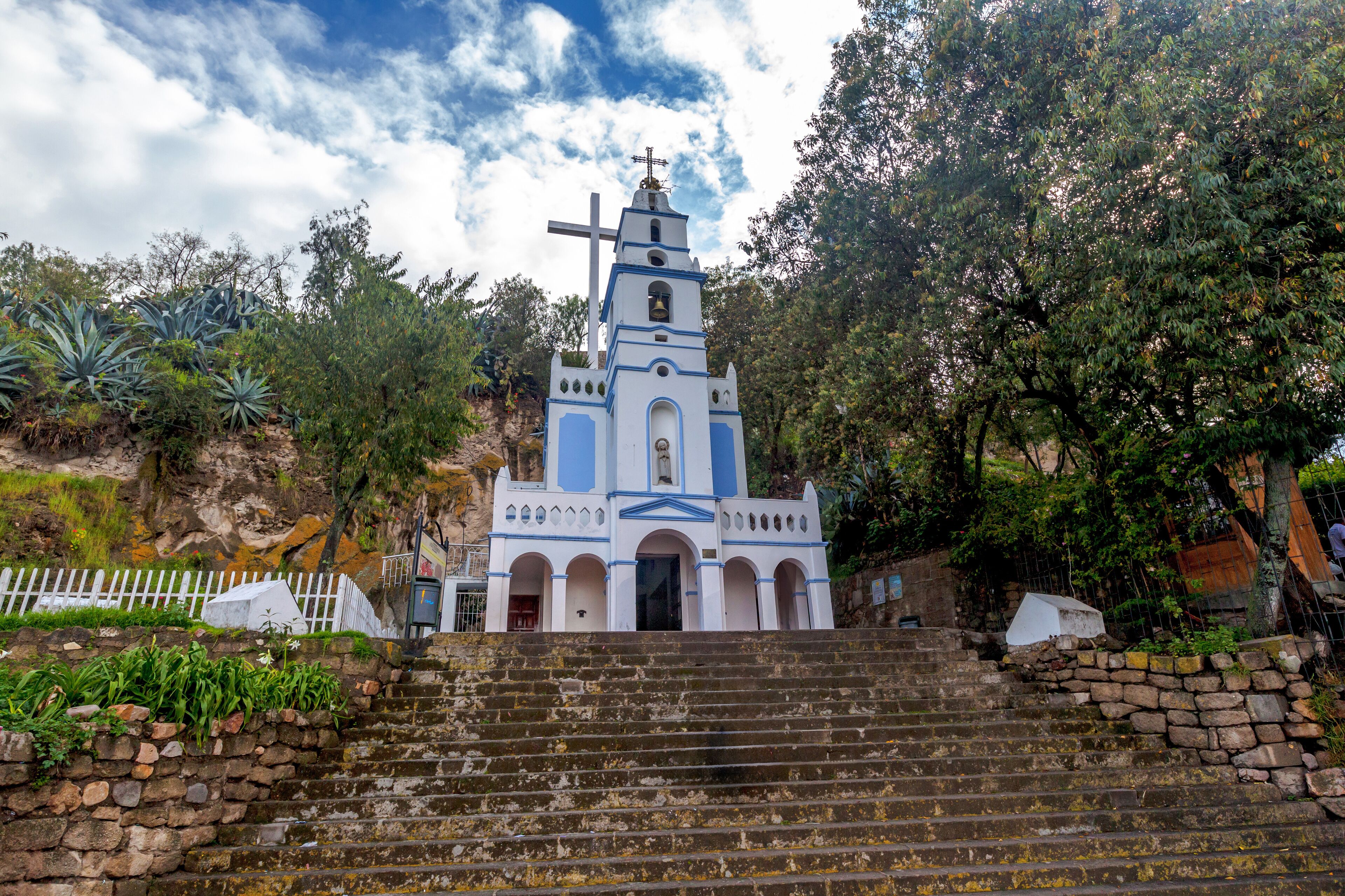 A white and blue church with a bell tower, surrounded by trees and a stone staircase leading up to it.