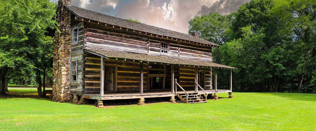 a brown log cabin in the park surrounded by lush green grass and trees with powerful clouds at McIntosh Reserve Park in Whitesburg Georgia