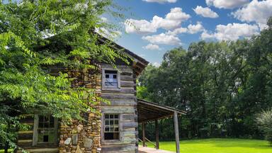 a brown log cabin in the park surrounded by lush green grass and trees with blue sky and clouds at McIntosh Reserve Park in Whitesburg Georgia