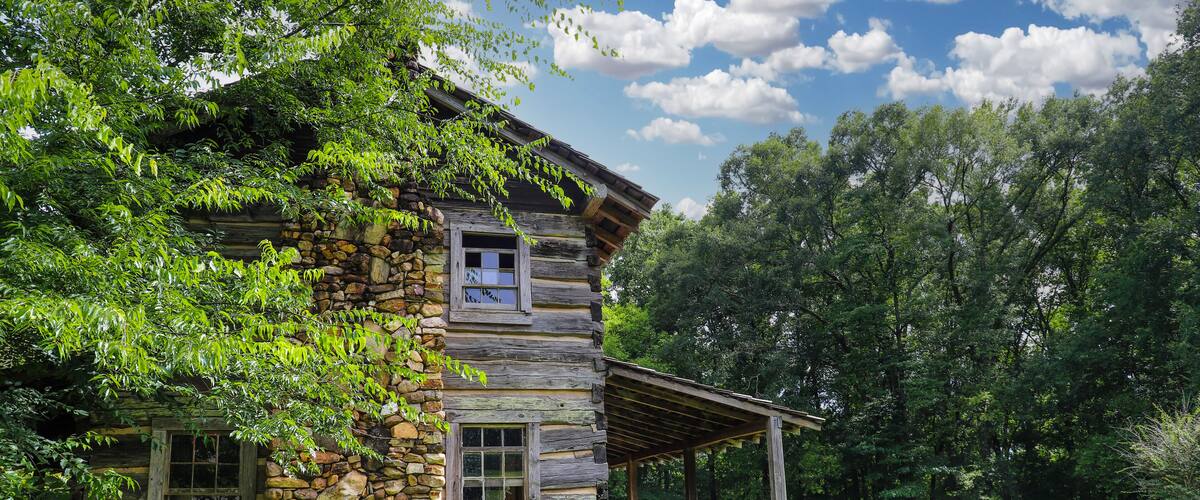 A rustic log cabin with a stone foundation, surrounded by lush greenery and a clear blue sky.