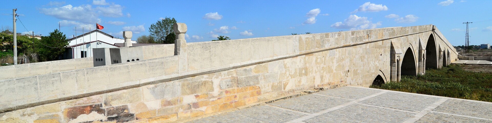 Un puente de piedra con arcos, una zona pavimentada y un pequeño edificio con techo rojo.