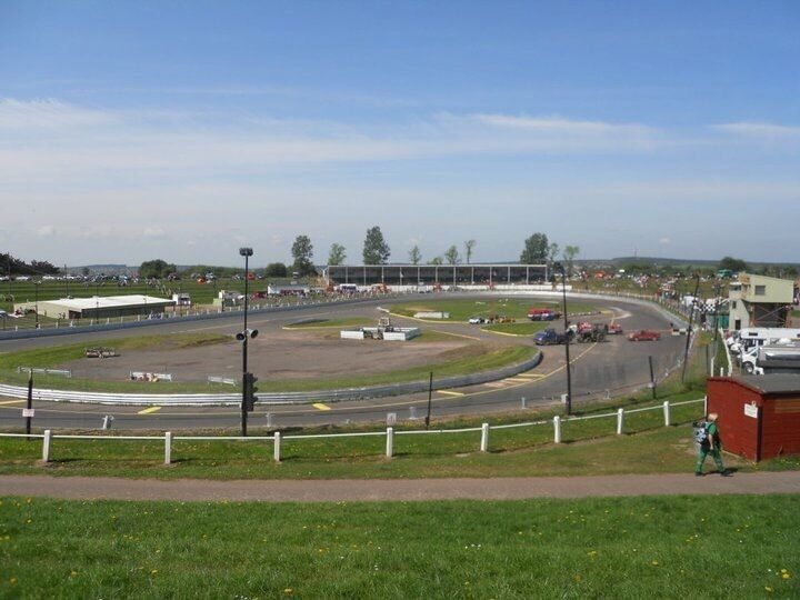 A race track with multiple lanes, spectator stands, and a grassy area with a fence.