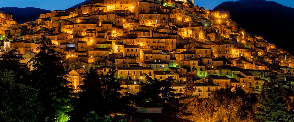 A densely packed hillside town at night, illuminated by streetlights and building lights, with a prominent church on top.