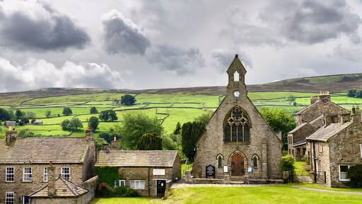 A stone church with a clock tower, surrounded by traditional stone houses and lush green fields.
