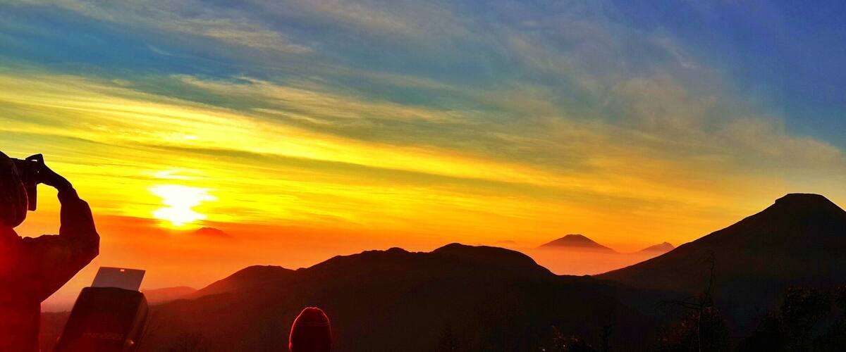 A sunset over a mountain range with a person taking a photo.