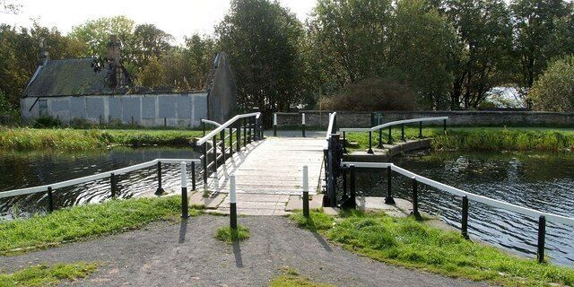 Ferrydyke Bascule Bridge. This bascule bridge, crossing the Forth and Clyde Canal, is shown from the other side at 939735. On its far side, a cycle route runs alongside the canal. The ruined building standing on the left was the bridgekeeper's cottage; see 1524165. Note also the low wall that runs alongside the cycle route on the right. From that wall, some sunken ruins can be seen on the other side; these are the remains of old stables: see 1524161. Between the ruined cottage and the stable wall, a path leads from the cycle route towards the former site of Donald's Quay, where a beacon still stands: 1493744. For a good summary of the history of this immediate area, see 961147.