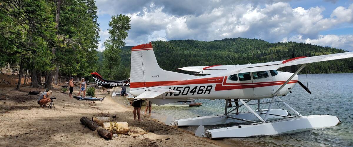 A seaplane on a sandy beach with water, trees, and a cloudy sky.