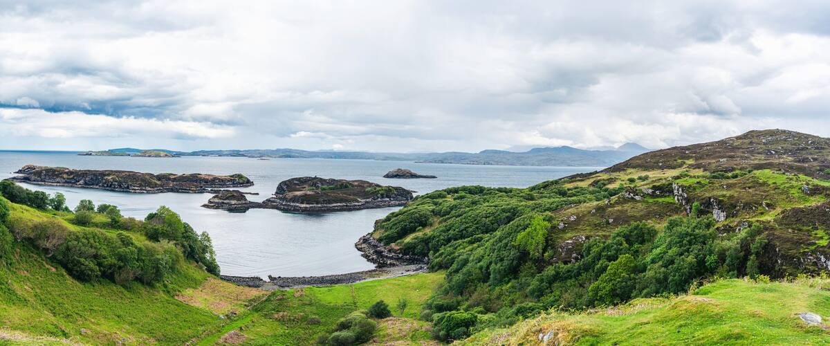 A coastal landscape with green hills, rocky outcrops, and a calm sea under a cloudy sky.