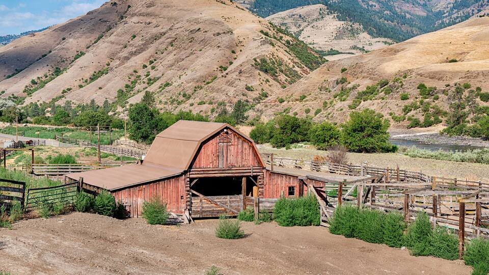 A rustic red barn with a metal roof, surrounded by a wooden fence and greenery, set against a backdrop of rolling hills and a clear sky.