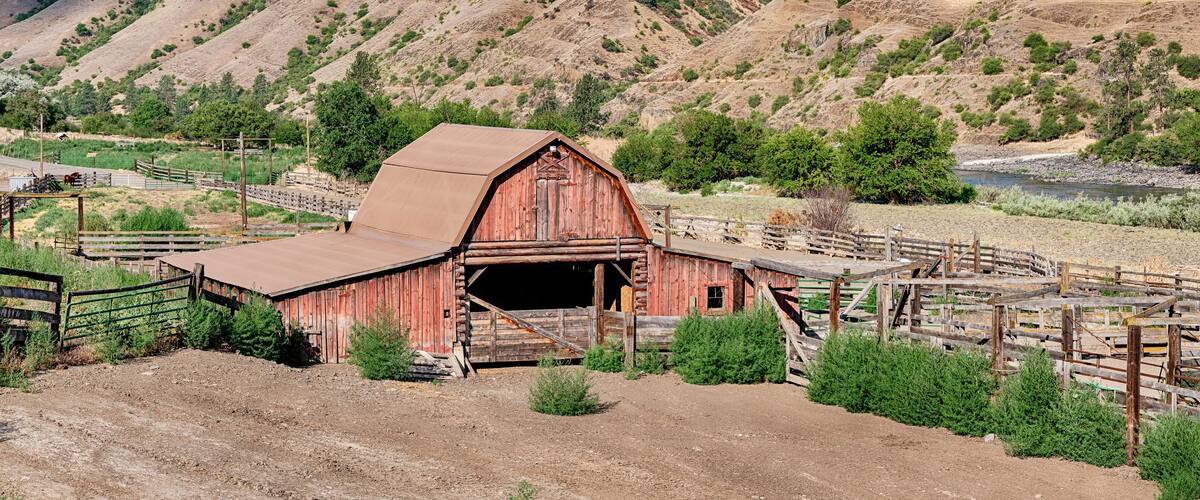 A rustic red barn with a metal roof, surrounded by a wooden fence and greenery, set against a backdrop of rolling hills and a clear sky.