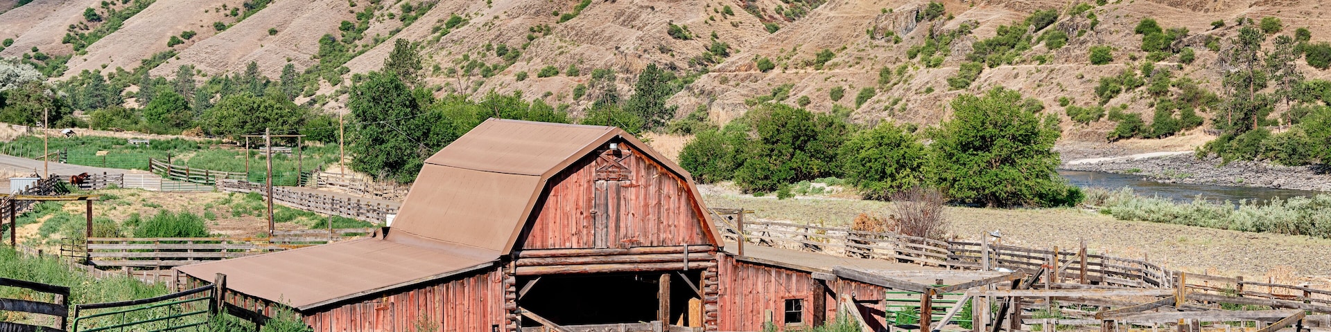 A rustic red barn with a metal roof, surrounded by a wooden fence and greenery, set against a backdrop of rolling hills and a clear sky.