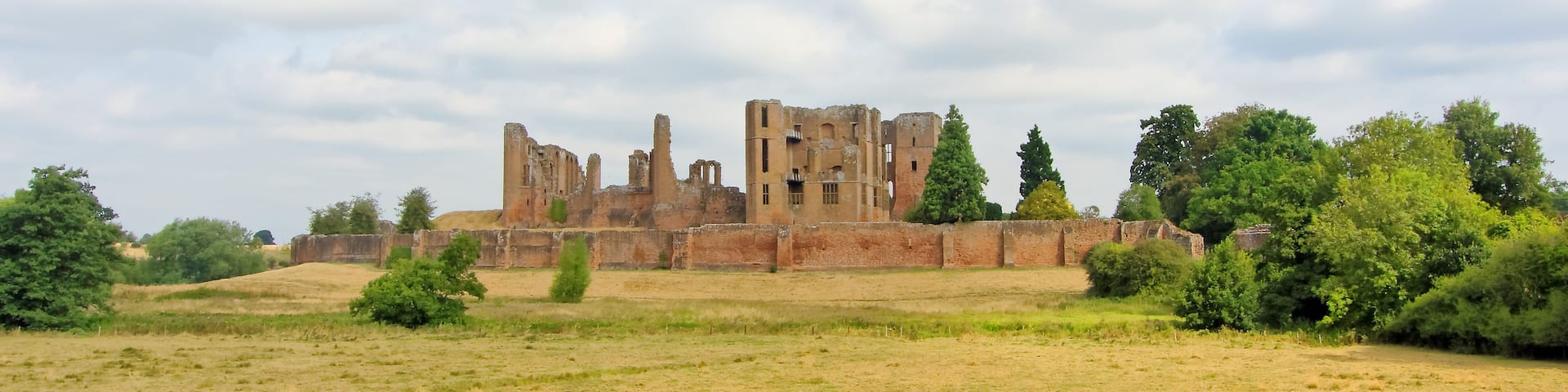 A historic castle ruin with a moat, surrounded by a grassy field and trees.