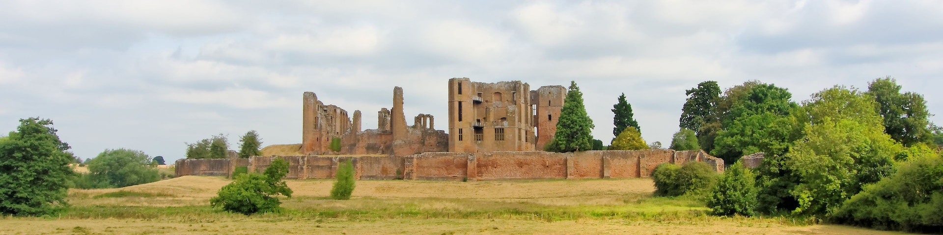 Ruinas de un castillo histórico con foso, rodeado por un campo cubierto de césped y árboles.