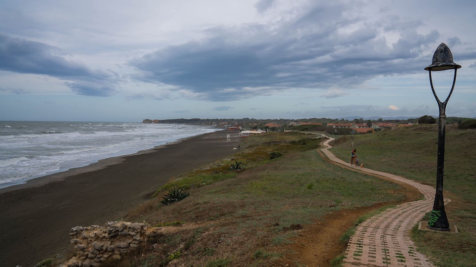A coastal path with a lamp post, beach, and distant buildings.